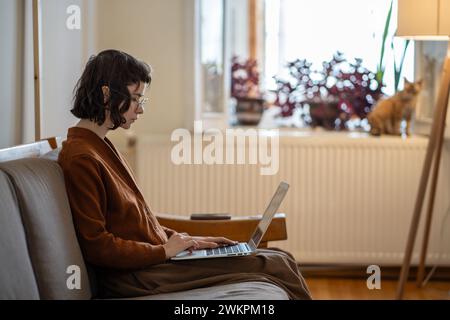 Studentessa seria e concentrata studiando a distanza online con un laptop in ginocchio seduto sul comodo divano di casa. Foto Stock