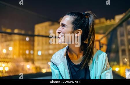 Ritratto di una giovane bruna allegra con coda di cavallo che guarda lontano in un ambiente urbano di notte. Donna felice con un sorriso genuino che esprime felicità Foto Stock