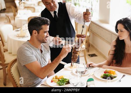 Coppia felice, appuntamento e cameriere con kebab per servire, cena raffinata o cena romantica al tavolo. Giovane uomo e donna con chef che tagliano spiedini di cibo Foto Stock