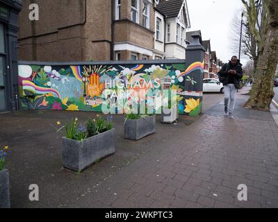 Un vivace murale di Street art dipinto su una parete esterna bianca sulla Avenue di Highams Park, Londra, Regno Unito, E4. Le opere d'arte raffigurano i monumenti locali e le scene dell'area di Highams Park, che fungono da opera d'arte pubblica concentrata sulla comunità nel cuore del quartiere. Situato nel London Borough of Waltham Forest, il murale contribuisce all'estetica suburbana della sfilata dello shopping vicino alla stazione ferroviaria di Highams Park Foto Stock