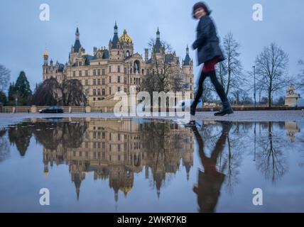 Schwerin, Germania. 22 febbraio 2024. Una donna cammina davanti a una pozzanghera in cui si riflette il castello di Schwerin. Con nebbia, nebbia e temperature di circa cinque gradi, l'inverno nel nord della Germania mostra il suo lato ostile. Crediti: Jens Büttner/dpa/Alamy Live News Foto Stock