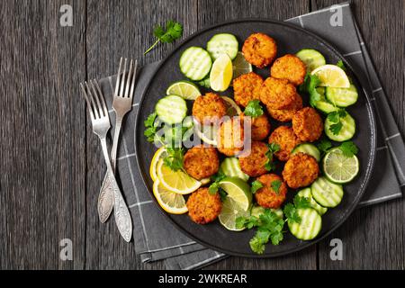 cotolette di riso fritte, tikki di riso, piatto indiano su piatto nero servito con cetriolo, limone, fette di lime e forchette su un tavolo di legno scuro, piatto, spalmabile Foto Stock