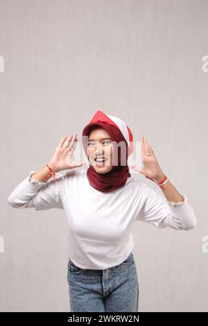 Giovane donna asiatica in hijab rosso e t-shirt bianca che celebra felicemente il giorno dell'indipendenza dell'Indonesia isolata su sfondo bianco, ritratto Foto Stock