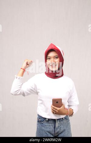 Una bella adolescente in un hijab che indossa una t-shirt bianca, regge un cellulare con la mano alzata. Concetto di indipendenza indonesiana. Foto Stock