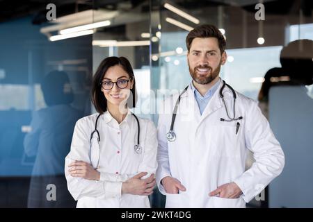 Due operatori sanitari, una donna e un uomo in camici bianchi con stetoscopi, sorridono con fiducia in un ambiente ospedaliero. Foto Stock