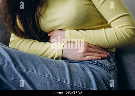 Primo piano di una giovane donna caucasica malata sconosciuta che soffre di mal di stomaco seduta sul divano a casa, tenendo la pancia, sentendosi addominale o mestruale pai Foto Stock