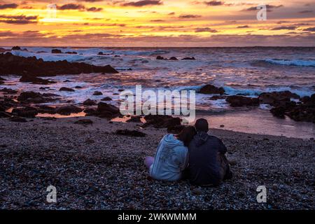 Giovane seduto sulla spiaggia al tramonto Foto Stock
