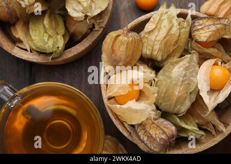 Frutti di physalis maturi con calici in ciotole e tazza di tè su tavola di legno, piatto Foto Stock