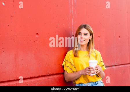 Una giovane donna gode di un momento di tranquillità con il suo caffè mattutino, appoggiandosi su un muro rosso testurizzato. La sua blusa gialla assolata e i jeans casual sk Foto Stock