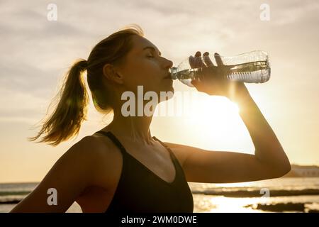 donna sportiva che beve l'acqua dalla bottiglia dopo un allenamento all'aperto al tramonto Foto Stock