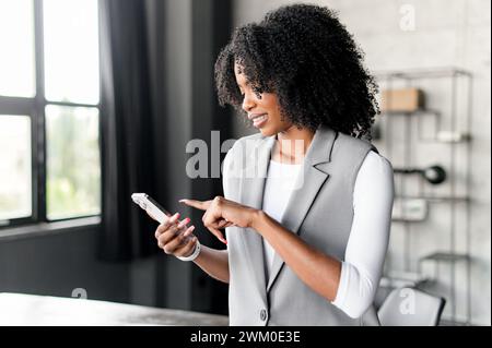 La stessa donna viene catturata in un sincero momento di gioia, navigando sul suo smartphone vicino alla finestra, inondata di luce naturale che mette in risalto la sua presenza sicura e tecnologicamente esperta. Foto Stock