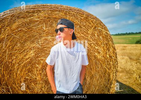 Ragazzo che soffia gomma da bolle e si appoggia sulla balla di paglia nel campo Foto Stock