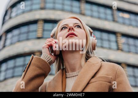 Donna contemplativa che ascolta la musica attraverso le cuffie wireless di fronte all'edificio Foto Stock