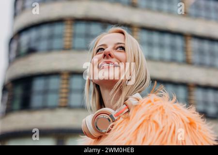 Donna bionda sorridente con cuffie wireless di fronte all'edificio Foto Stock