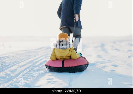 Padre che tira figlio che si trova sul tubo gonfiabile in inverno Foto Stock