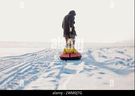 Padre maturo che tira figlio che siede sul tubo gonfiabile nella neve Foto Stock