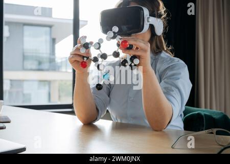 Scienziato che indossa cuffie per realtà virtuale e tiene la struttura molecolare alla scrivania Foto Stock