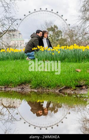 Londra, Regno Unito. 23 febbraio 2024. Un paio di ourists si fermano per scattare un selfie tra i fiori, usando un supporto per telefono, mentre l'occhio di Londra si riflette in una delle pozzanghere di acqua stagnante lasciate dopo il maltempo degli ultimi giorni - primi segni di primavera quando i narcisi iniziano a uscire, c'è una breve pausa sotto la pioggia e il sole tenta di uscire a St James Park. Crediti: Guy Bell/Alamy Live News Foto Stock