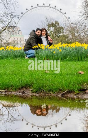 Londra, Regno Unito. 23 febbraio 2024. Un paio di ourists si fermano per scattare un selfie tra i fiori, usando un supporto per telefono, mentre l'occhio di Londra si riflette in una delle pozzanghere di acqua stagnante lasciate dopo il maltempo degli ultimi giorni - primi segni di primavera quando i narcisi iniziano a uscire, c'è una breve pausa sotto la pioggia e il sole tenta di uscire a St James Park. Crediti: Guy Bell/Alamy Live News Foto Stock