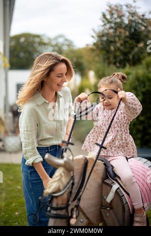 Ragazza felice che gioca sul cavallo giocattolo vicino alla madre in giardino Foto Stock