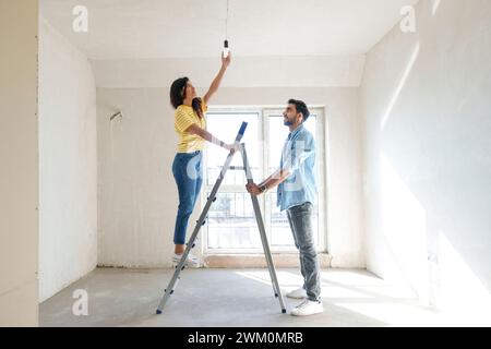 Giovane coppia che usa la scala e installa la lampadina nella nuova casa Foto Stock