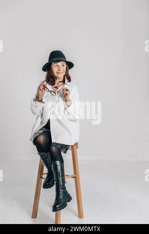 Donna anziana alla moda seduta sullo sgabello di fronte al muro bianco Foto Stock