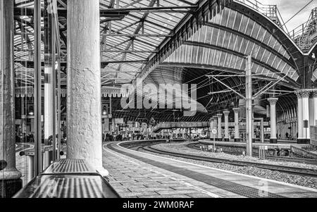 Uno storico arco metallico copre l'ingresso di una stazione ferroviaria. Un treno e i passeggeri stanno ai binari e una colonna è in primo piano. Foto Stock