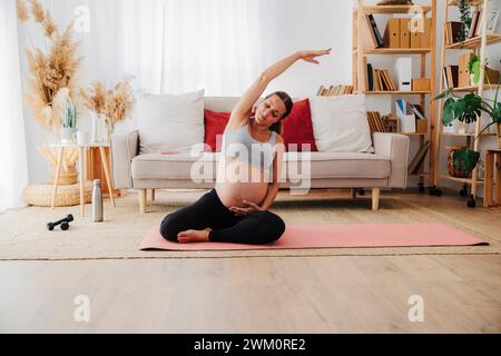 Donna incinta che si allunga la mano praticando yoga su un tappetino in salotto di casa Foto Stock