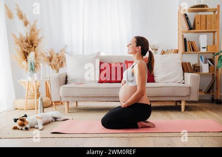 Donna incinta inginocchiata con le mani sullo stomaco che meditava vicino al cane in salotto Foto Stock