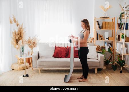 Donna incinta che rotola il tappetino yoga nel soggiorno di casa Foto Stock