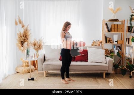 Donna incinta che rotola il tappetino yoga in soggiorno Foto Stock