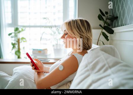 Donna che utilizza uno smartphone con sensore di glucosio sul braccio a casa Foto Stock