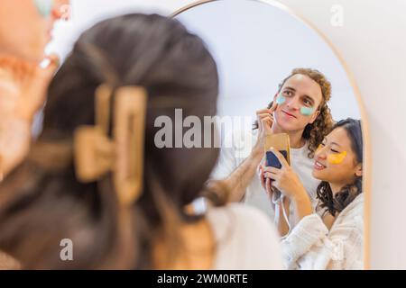 Coppia sorridente che fa la cura della pelle e scatta foto davanti allo specchio a casa Foto Stock
