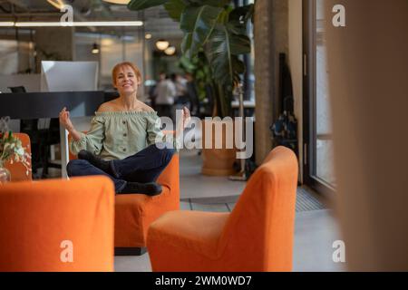 Donna d'affari matura sorridente seduta su una sedia arancione in ufficio Foto Stock