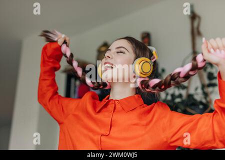 Giovane donna felice che tiene le trecce e ascolta la musica a casa Foto Stock