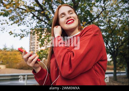 Donna sorridente rossa che regola le cuffie intrauricolari cablate nel parco autunnale Foto Stock