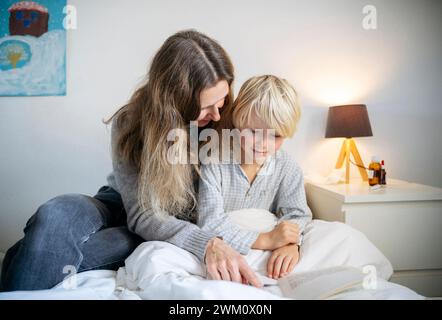 Madre che legge un libro a un figlio seduto sul letto a casa Foto Stock