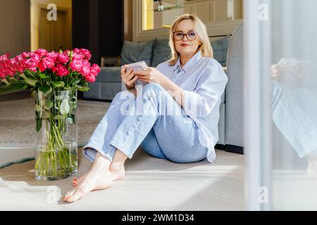 Donna matura di 55 anni donna bionda ben curata con occhiali, vestita con jeans e camicia blu di lino, seduta sul pavimento del soggiorno, a leggere una Foto Stock