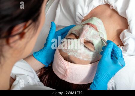 L'estetista applica la maschera facciale verde a base di erbe sul viso del cliente. Donna di mezza età che riceve trattamenti termali in un salone di bellezza. Foto Stock