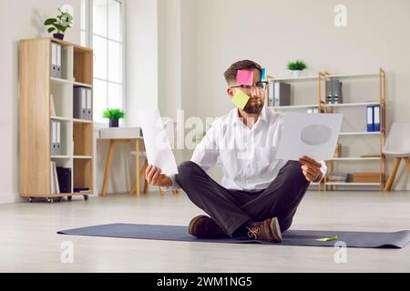 Uomo divertente seduto sul pavimento su un tappetino yoga con note sul viso e leggendo documenti in ufficio. Foto Stock