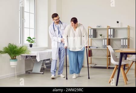 Giovane medico allegro e amichevole che aiuta una donna grassa a camminare con le stampelle in clinica. Foto Stock