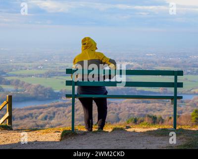 donna seduta sulla panchina in cima alla collina avvolta da un caldo cappotto con cappuccio sollevato per proteggersi dal vento freddo Foto Stock