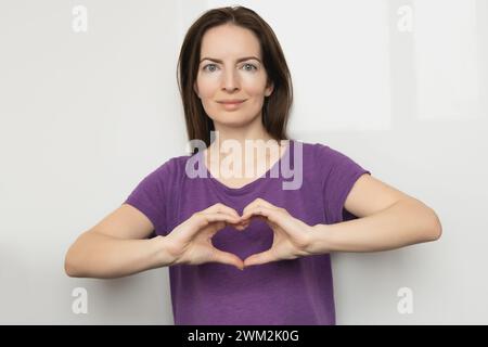 Ispira l'inclusione. Donna che tiene le mani a forma di cuore e le tiene davanti a sé, t-shirt viola vestita. Giornata internazionale delle donne Foto Stock