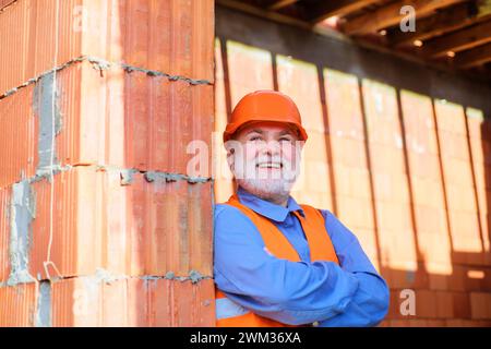 Lavoratore sorridente in giubbotto di sicurezza e casco sul lavoro. Architetto barbato costruttore di elmetto protettivo. Ristrutturazione di edifici e abitazioni Foto Stock