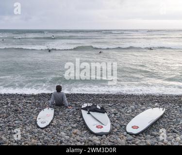 Surfista accanto a tre tavole da surf su una spiaggia con ciottoli, pronto ad entrare in acqua a Lima, Perù Foto Stock