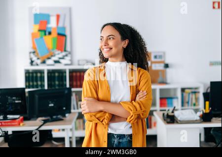 Positivo sicuro di sé bella donna d'affari ispanica dai capelli ricci con camicia arancione, business coach, manager, in piedi in un ufficio moderno con le braccia incrociate, guardando lontano e sorridendo Foto Stock
