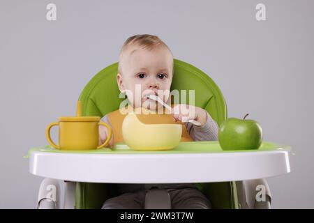 Piccolo bambino carino che mangia cibo sano in seggiolone su sfondo grigio Foto Stock