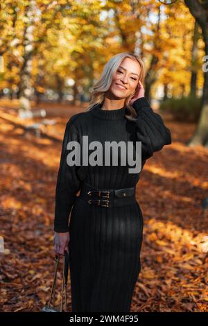 Bella giovane donna felice in abiti alla moda con un elegante abito nero lavorato a maglia con una borsa cammina in un parco d'autunno dorato Foto Stock