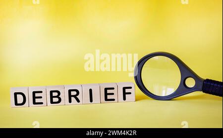 Cubi in legno con testo di debrief su sfondo giallo accanto a una lente d'ingrandimento Foto Stock