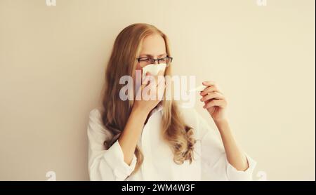Donna caucasica stanca malata dipendente starnuti soffiare naso utilizzando il tessuto e misurare la temperatura corporea guardando il termometro a casa Foto Stock